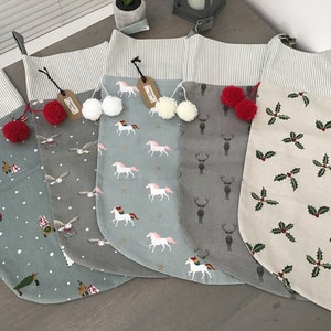 May include: Five Christmas stockings with different patterns. The stockings are made of a light-coloured fabric with a white stripe at the top. The patterns include a unicorn, a reindeer, a snowman, an owl, and holly leaves. Each stocking has a pom-pom at the top.