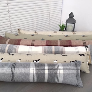 May include: A stack of five doorstop cushions in various patterns. The bottom cushion is grey and white plaid, the next is beige with black dog silhouettes, the next is red and white plaid, the next is green with brown pheasant silhouettes, and the top is beige with a brown pheasant silhouette.