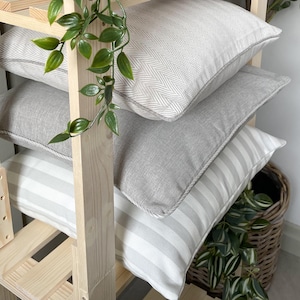 May include: Four decorative throw pillows stacked on a wooden shelf. The pillows feature various patterns, including herringbone, solid gray, and striped designs. Green leafy vines are draped around the shelf, adding a natural touch.