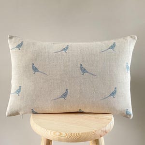 Puede incluir: Un cojín rectangular beige con un patrón repetido de ilustraciones de faisanes azules. El cojín está colocado sobre un taburete de madera clara contra un fondo neutro. Un accesorio decorativo para el hogar.