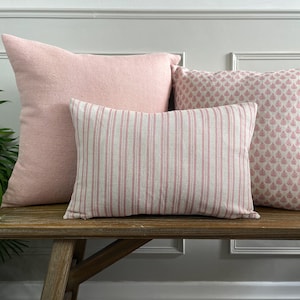 May include: Three pink cushions on a wooden bench. The cushions are made of different fabrics and have different patterns. The cushion on the left is a solid pink, the middle cushion has pink and white stripes, and the cushion on the right has a pink floral pattern.
