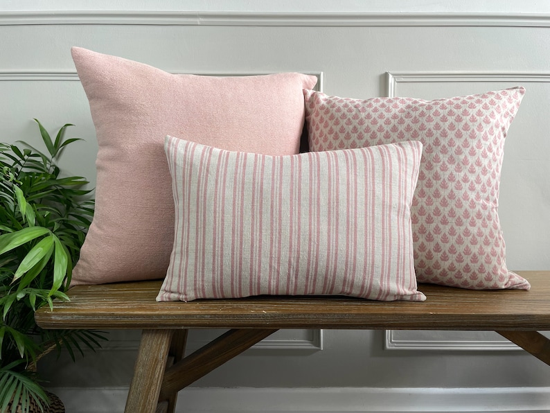 May include: Three pink cushions on a wooden bench. The cushions are made of different fabrics and have different patterns. The cushion on the left is a solid pink, the middle cushion has pink and white stripes, and the cushion on the right has a pink floral pattern.
