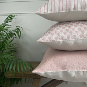 May include: Three pink and white patterned throw pillows stacked on a wooden bench. The top pillow has pink stripes, the middle pillow has a pink floral pattern, and the bottom pillow is a solid pink colour.
