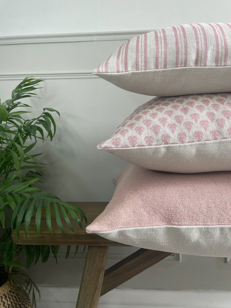 May include: Three pink and white patterned throw pillows stacked on a wooden bench. The top pillow has pink stripes, the middle pillow has a pink floral pattern, and the bottom pillow is a solid pink colour.