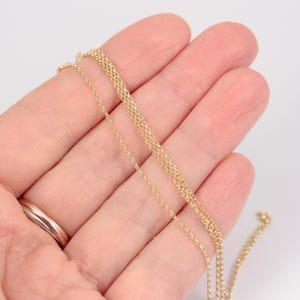 K&ouml;nnte beinhalten: Zarte goldene Halskette mit feinem, strukturiertem Design. Die Halskette wird vor einem hellen Hintergrund pr&auml;sentiert, der ihre subtilen Details und ihre warme Farbe hervorhebt. Die Kette wird in einer Hand gehalten, um ihre L&auml;nge und zarte Konstruktion zu zeigen.