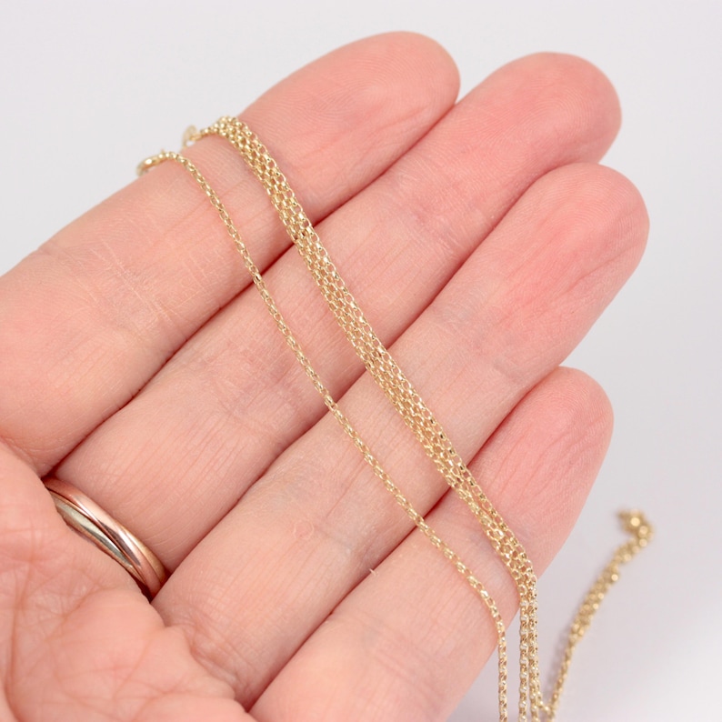 K&ouml;nnte beinhalten: Zarte goldene Halskette mit feinem, strukturiertem Design. Die Halskette wird vor einem hellen Hintergrund pr&auml;sentiert, der ihre subtilen Details und ihre warme Farbe hervorhebt. Die Kette wird in einer Hand gehalten, um ihre L&auml;nge und zarte Konstruktion zu zeigen.