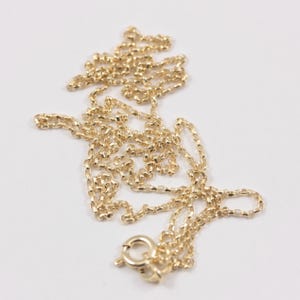 K&ouml;nnte beinhalten: Eine zarte, goldfarbene Halskette mit kleinem Verschluss. Die Kette besteht aus kleinen, verbundenen Gliedern, die ein strukturiertes Aussehen erzeugen. Die Halskette wird auf einer wei&szlig;en Oberfl&auml;che pr&auml;sentiert, um ihre Farbe und ihr Design hervorzuheben.