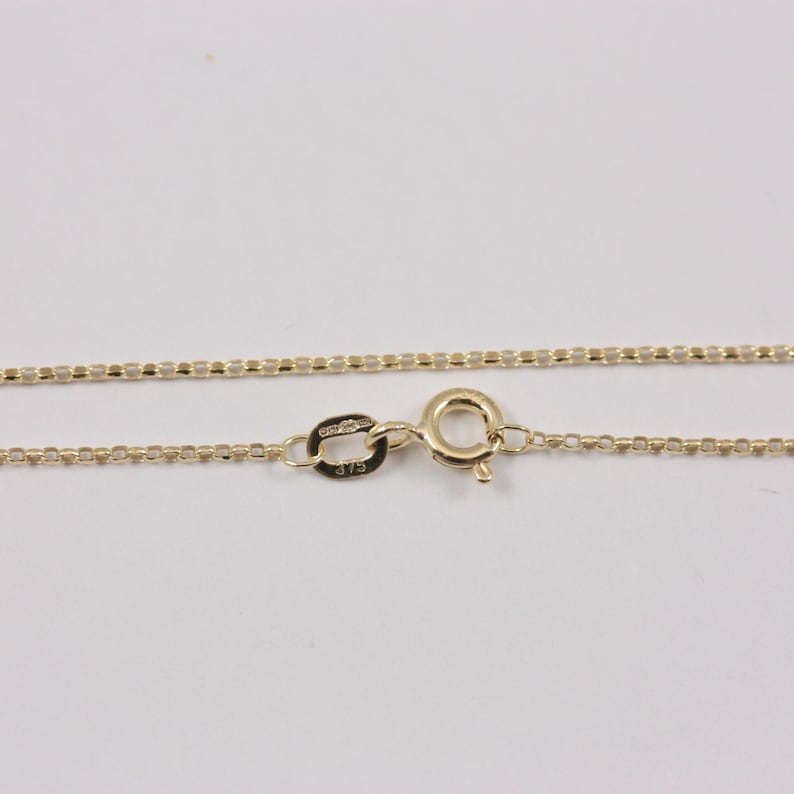 K&ouml;nnte beinhalten: Eine filigrane Goldkette mit einem Box-Chain-Design. Die Halskette hat einen Karabinerverschluss und ein kleines Etikett mit der Pr&auml;gung "375". Die Kette wird vor einem wei&szlig;en Hintergrund gezeigt.