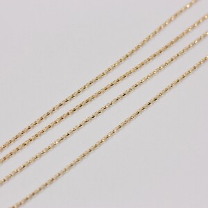 K&ouml;nnte beinhalten: Vier zarte Goldketten mit einem strukturierten, facettierten Design. Die Ketten sind diagonal auf einem wei&szlig;en Hintergrund angeordnet und bilden ein Muster aus parallelen Linien. Die Goldfarbe ist warm und hell.
