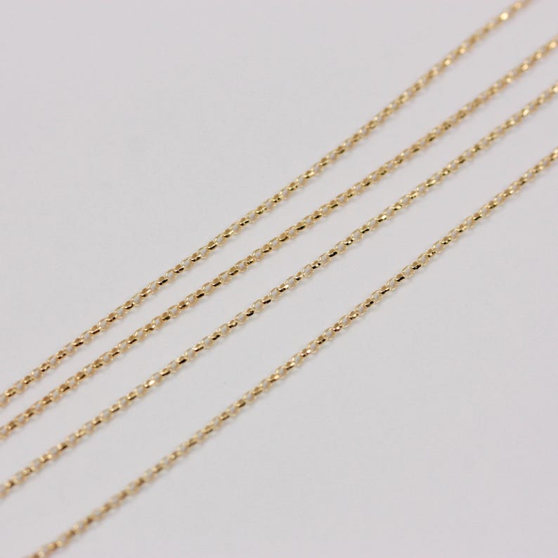 K&ouml;nnte beinhalten: Vier zarte Goldketten mit einem strukturierten, facettierten Design. Die Ketten sind diagonal auf einem wei&szlig;en Hintergrund angeordnet und bilden ein Muster aus parallelen Linien. Die Goldfarbe ist warm und hell.