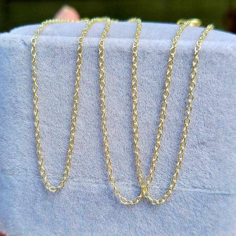 May include: Several delicate gold-colored chain necklaces with small, oval links are displayed against a soft, light blue background. The chains are draped, showcasing their length and design. The chains are likely for jewelry.