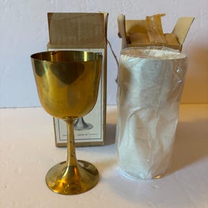 May include: A vintage gold-colored metal goblet with a wide bowl and slender stem, next to a cardboard box and a roll of white paper. The goblet has a shiny finish. The box has a printed image of the goblet.