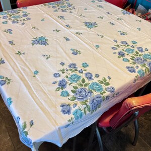 May include: A white tablecloth with a floral pattern of blue roses and green leaves. The tablecloth is rectangular and covers a square table. Red chairs surround the table.