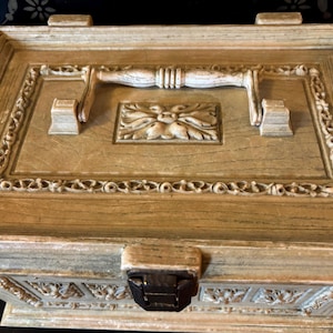 May include: A wooden box with a carved design and a white painted finish. The box has a hinged lid with a decorative handle and a latch.