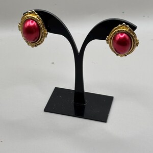 May include: A pair of vintage clip-on earrings. Each earring features a large, oval, red faux pearl center, surrounded by a gold-toned, ornate frame. The earrings are displayed on a black earring stand against a white background.