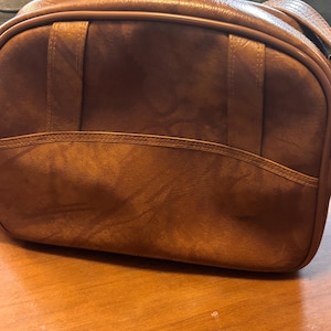 May include: A rectangular brown shoulder bag with a textured finish. The bag features a horizontal seam across the middle, vertical stitched accents on the front panel, and a matching brown piping trim along the edges of the main compartment.