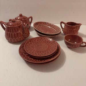 May include: A terracotta-colored ceramic tea set with a teapot, plates, a creamer, and a teacup. The plates have a sunflower design. The teapot and creamer have handles. The set is suitable for a dollhouse or decorative display.