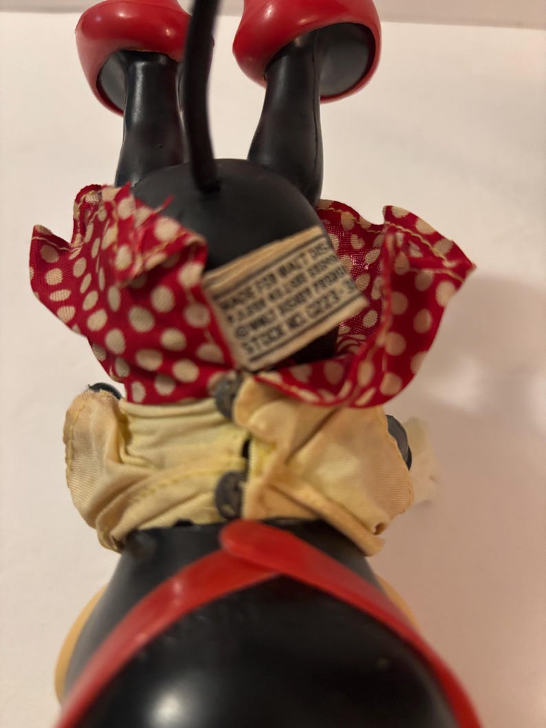 May include: Vintage Minnie Mouse figurine, with a black body, red shoes, and a red and white polka dot skirt. The figurine has a yellow top and red suspenders. A tag is attached to the back.