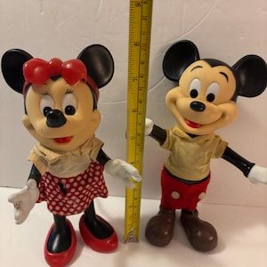 May include: Two vintage Mickey Mouse and Minnie Mouse figurines. Mickey wears red shorts and a yellow shirt. Minnie wears a red polka dot skirt and a red bow. A ruler shows the figurines are approximately 20 cm tall.