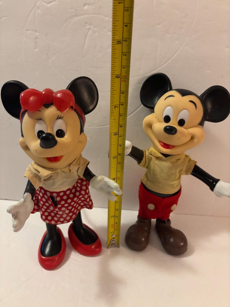 May include: Two vintage Mickey Mouse and Minnie Mouse figurines. Mickey wears red shorts and a yellow shirt. Minnie wears a red polka dot skirt and a red bow. A ruler shows the figurines are approximately 20 cm tall.