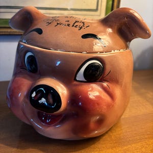 May include: A vintage ceramic pig-shaped cookie jar with a winking eye and a smiling expression. The jar is primarily brown with red accents on the cheeks and snout. Text on the lid reads "Go ahead, take a pig of yourself!"