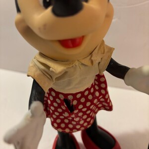 May include: A Minnie Mouse figurine with black features, wearing a yellow top and a red skirt with white polka dots. The shoes are red, and the gloves are white. The figurine has a red bow.