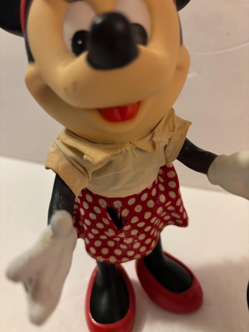 May include: A Minnie Mouse figurine with black features, wearing a yellow top and a red skirt with white polka dots. The shoes are red, and the gloves are white. The figurine has a red bow.
