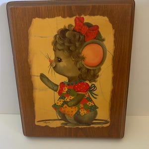 May include: A wooden plaque with a painted image of a cartoon mouse wearing a red and white polka dot dress and a red bow. The mouse is standing on a yellow background with a brown wooden frame.