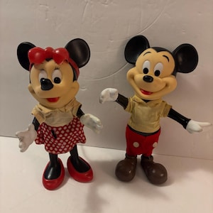 May include: Two vintage Mickey and Minnie Mouse figurines. Mickey wears red shorts, brown shoes, and a yellow shirt. Minnie wears a red polka dot skirt, red shoes, and a red bow. Both have black ears and white gloves.