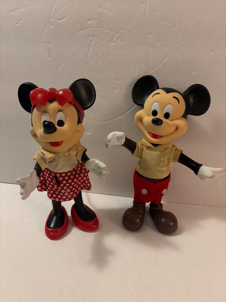May include: Two vintage Mickey and Minnie Mouse figurines. Mickey wears red shorts, brown shoes, and a yellow shirt. Minnie wears a red polka dot skirt, red shoes, and a red bow. Both have black ears and white gloves.