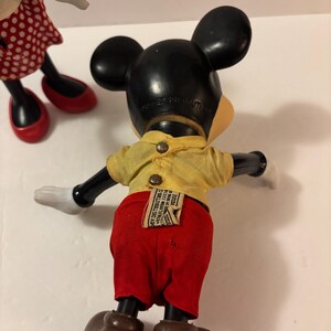 May include: Vintage Mickey Mouse doll with black ears, a yellow shirt, red shorts, and brown shoes. The doll has white gloves and a tag on the back. Another doll with a red polka dot skirt and red shoes is partially visible.