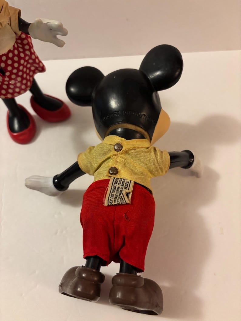 May include: Vintage Mickey Mouse doll with black ears, a yellow shirt, red shorts, and brown shoes. The doll has white gloves and a tag on the back. Another doll with a red polka dot skirt and red shoes is partially visible.