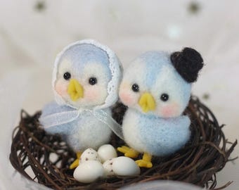 May include: Two blue felt birds with yellow beaks and black eyes are sitting in a brown twig nest. The bird on the left is wearing a white lace bonnet. The bird on the right is wearing a black felt top hat. There are five white speckled eggs in the nest.