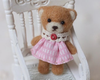 May include: A miniature brown teddy bear in a pink and white dress and a white lace collar with a red button. The bear sits on a small white wooden chair with a decorative back.