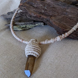 May include: A white cord necklace with a copper wire accent and a brown crystal pendant. The necklace is wrapped around a piece of driftwood.