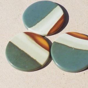 Może przedstawiać: Trzy okrągłe ceramiczne podstawki. Każda podstawka podzielona jest na trzy sekcje: morski, biały i pomarańczowo-brązowy pas. Podstawki mają błyszczące wykończenie i są ułożone na jasnobrązowej powierzchni.