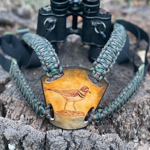 May include: A green and brown camouflage paracord binocular harness with a leather chest strap featuring a hand-painted image of a killdeer bird.