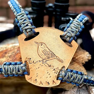 May include: A tan leather binocular harness with a detailed bird engraving. The harness features blue and camouflage paracord straps. The background shows a pair of binoculars.