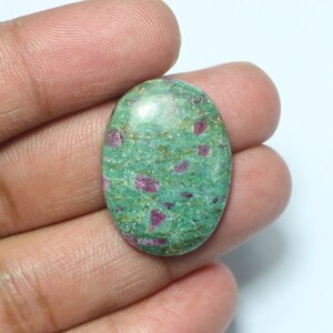 May include: An oval gemstone with a green base and red inclusions. The stone has a polished surface, and the red inclusions are irregularly shaped. The gemstone is held in a hand.