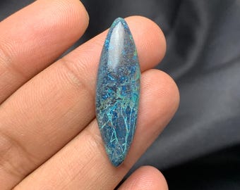 Può includere: Una gemma lucida e allungata con una punta affilata. La pietra presenta una miscela di tonalità blu e verdi con intricati motivi a forma di venature. La gemma è tenuta in mano su uno sfondo scuro.