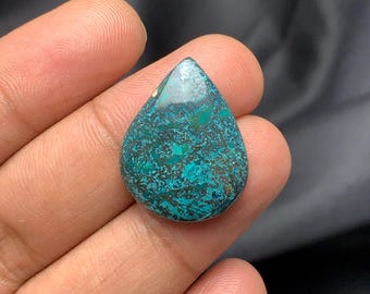 Può includere: Una gemma a forma di goccia con una miscela di colori verde acqua, turchese e marrone. La superficie lucida mostra un motivo screziato, con aree di verde acqua intenso e tonalità turchesi più chiare. La pietra è tenuta in una mano.