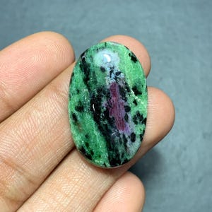Puede incluir: Una piedra preciosa cabujón de forma ovalada que presenta una base verde vibrante con prominentes inclusiones minerales negras y una mancha central distintiva de material cristalino de color magenta intenso, pulida y lisa.