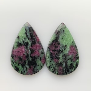 Peut inclure: Deux pierres précieuses de rubis zoïsite en forme de goutte. Les pierres présentent un mélange de couleurs vertes, noires et magenta. Les pierres précieuses sont polies et ont une surface lisse. Le fond est blanc.