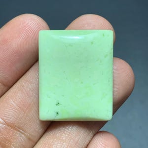 以下が含まれることがあります： 滑らかで平らな表面を持つ、鮮やかなライトグリーンの長方形の研磨された宝石。不透明な石の全体に繊細な斑点模様があり、左下隅付近に小さなダークグリーンのインクルージョンが見られます。