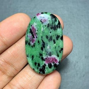 Può includere: Una pietra preziosa a cabochon di forma ovale con una base in zoisite verde brillante, caratterizzata da inclusioni distinte di rubino magenta intenso e cristalli di orneblenda neri sparsi, lucidata con una finitura liscia e riflettente.