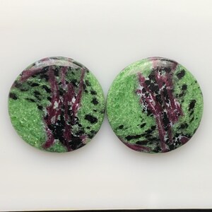 Könnte beinhalten: Zwei runde Zoisit-Edelsteine. Die Steine haben eine grüne Grundfarbe mit schwarzen und kastanienbraunen Einschlüssen. Die Edelsteine sind poliert und haben eine glatte, glänzende Oberfläche. Die Steine sind vor einem weißen Hintergrund platziert.