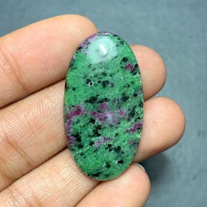 May include: An oval-shaped cabochon gemstone featuring a vibrant green base color with scattered black mineral inclusions and distinct patches of deep magenta-red throughout the polished surface, showcasing a smooth and reflective texture.