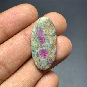 May include: An oval-shaped cabochon gemstone featuring a mottled surface with vibrant magenta-pink inclusions set against a matrix of pale green, teal, and earthy tan mineral deposits, displaying a polished, smooth, and rounded texture.