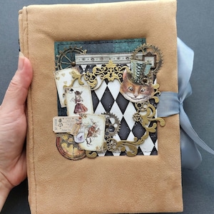 Op de afbeelding: Een notitieboek met een beige suède-achtige textuur en een centraal paneel met een zwart-wit ruitpatroon. De kaft is versierd met metalen tandwielen, speelkaarten, een illustratie van een kat met een hoge hoed en een lichtblauw lint.