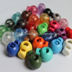 May include: Assortment of colourful plastic toggle clasps, also known as cord locks, in various colours including blue, green, red, orange, yellow, and white.  These are used for adjusting the length of cords, drawstrings, and straps.